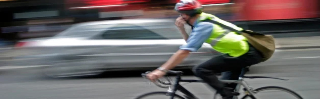 A man wearing a helmet and Hi Vis jacket is riding a bike. A silver car is passing on the opposite side of the road. 