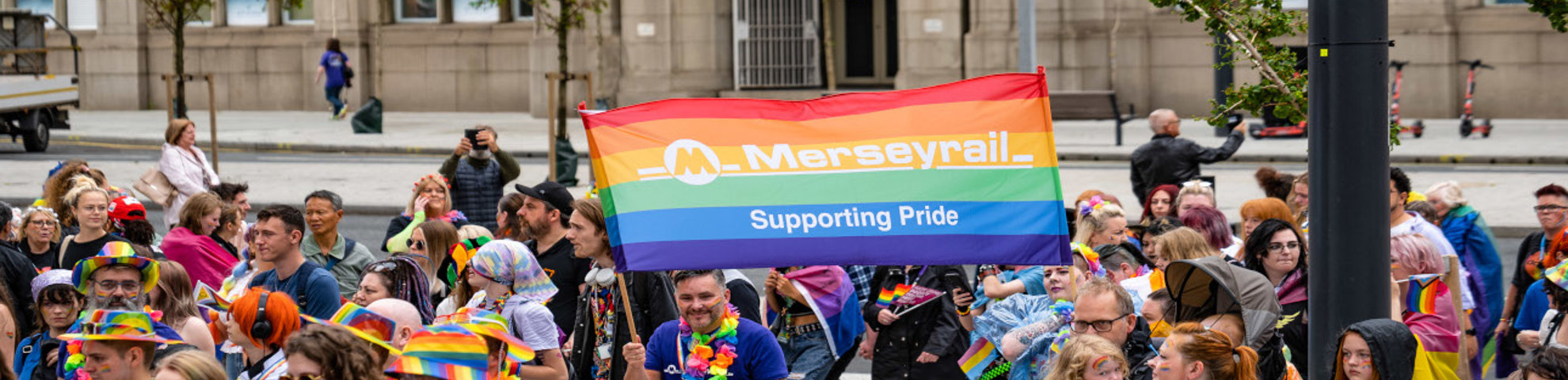 Merseyrail staff attending pride, with two members of staff holding a large banner of the rainbow-coloured flag with the Merseyrail logo on 