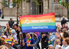Merseyrail staff attending pride, with two members of staff holding a large banner of the rainbow-coloured flag with the Merseyrail logo on