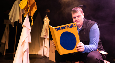 The Boy King play. A man is reading a book on stage.